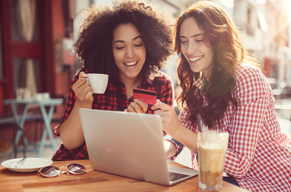 To piger drikker kaffe og ser på et Bank Norwegian-kort mens computeren står tændt