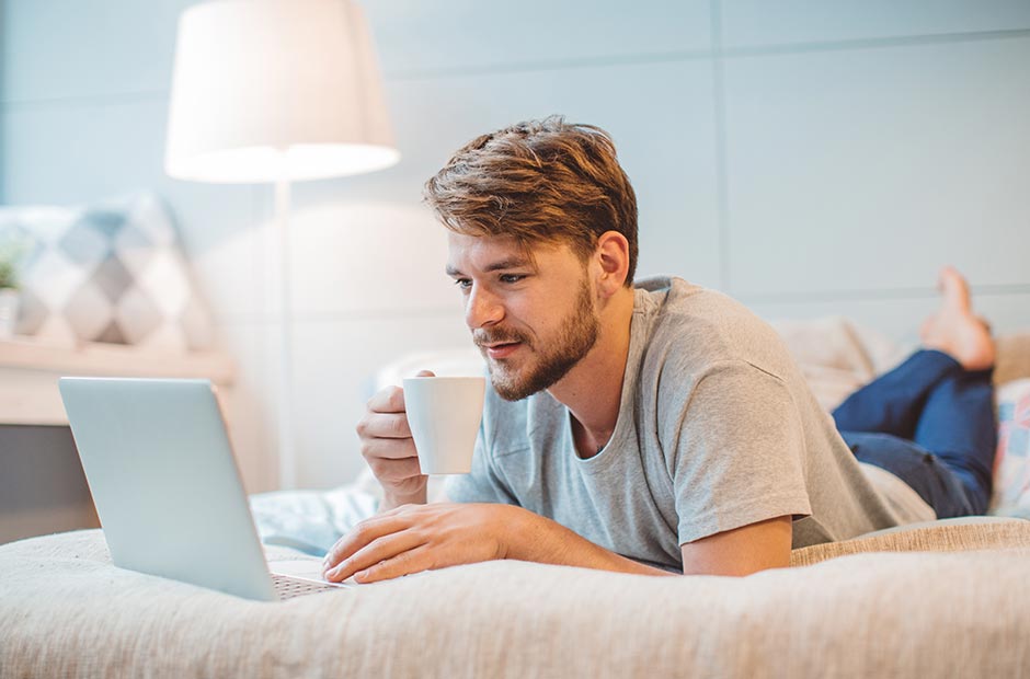 Mand ligger I sengen og drikker en varm drik mens han læser emails på sin computer